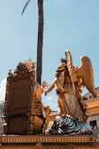 An ornate golden sculpture featuring an angel with large wings kneeling beside an elaborately carved throne. The scene is set against a clear blue sky with a tall palm tree in the background. People in the background are watching the display from a balcony.