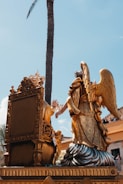 An ornate golden sculpture featuring an angel with large wings kneeling beside an elaborately carved throne. The scene is set against a clear blue sky with a tall palm tree in the background. People in the background are watching the display from a balcony.
