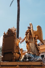 An ornate golden sculpture featuring an angel with large wings kneeling beside an elaborately carved throne. The scene is set against a clear blue sky with a tall palm tree in the background. People in the background are watching the display from a balcony.