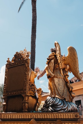An ornate golden sculpture featuring an angel with large wings kneeling beside an elaborately carved throne. The scene is set against a clear blue sky with a tall palm tree in the background. People in the background are watching the display from a balcony.