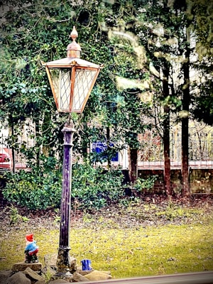 A vintage-style street lamp with a copper top stands in a garden setting surrounded by lush greenery. At the base of the lamp, a small garden gnome is visible, positioned among stones on a grassy area. Behind the lamp, dense trees and a wrought iron fence are seen, partially obscuring objects in the background.