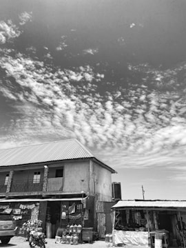A two-story building with a corrugated metal roof is pictured, surrounded by a few small shops or stalls. The sky is partly cloudy with scattered, fluffy clouds in the background. Several items, possibly goods for sale, are displayed in front of the stalls.