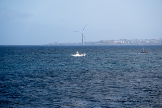 A vast ocean with a small white boat and sailboat on the water. In the background, a large white wind turbine stands near a distant coastline, which is faintly visible along with some buildings.