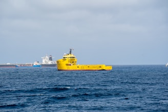A bright yellow ship is prominently positioned on the calm ocean waters, with a clear blue sky overhead. In the background, there are multiple ships or vessels, including a large white ship and a smaller blue one near a port or industrial area.