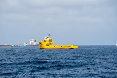 A bright yellow ship is prominently positioned on the calm ocean waters, with a clear blue sky overhead. In the background, there are multiple ships or vessels, including a large white ship and a smaller blue one near a port or industrial area.