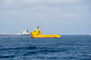 A bright yellow ship is prominently positioned on the calm ocean waters, with a clear blue sky overhead. In the background, there are multiple ships or vessels, including a large white ship and a smaller blue one near a port or industrial area.