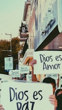 A group of people is holding up signs with messages in Spanish, such as 'Dios es Amor' and 'Dios es Paz'. They stand on a city street near a building with balconies, surrounded by urban elements like street lamps and trees.
