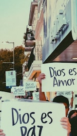 A group of people is holding up signs with messages in Spanish, such as 'Dios es Amor' and 'Dios es Paz'. They stand on a city street near a building with balconies, surrounded by urban elements like street lamps and trees.