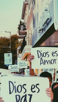 A group of people is holding up signs with messages in Spanish, such as 'Dios es Amor' and 'Dios es Paz'. They stand on a city street near a building with balconies, surrounded by urban elements like street lamps and trees.