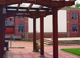 An outdoor space with wooden pergolas framing a courtyard. The ground is paved with bricks, and a mural depicting people in windows is painted on a building facade in the background. Vibrant colors on fabric decorations are visible alongside red brick walls.
