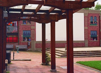 An outdoor space with wooden pergolas framing a courtyard. The ground is paved with bricks, and a mural depicting people in windows is painted on a building facade in the background. Vibrant colors on fabric decorations are visible alongside red brick walls.
