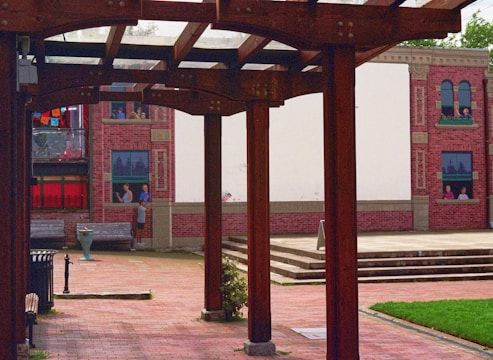 An outdoor space with wooden pergolas framing a courtyard. The ground is paved with bricks, and a mural depicting people in windows is painted on a building facade in the background. Vibrant colors on fabric decorations are visible alongside red brick walls.