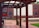 An outdoor space with wooden pergolas framing a courtyard. The ground is paved with bricks, and a mural depicting people in windows is painted on a building facade in the background. Vibrant colors on fabric decorations are visible alongside red brick walls.