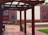 An outdoor space with wooden pergolas framing a courtyard. The ground is paved with bricks, and a mural depicting people in windows is painted on a building facade in the background. Vibrant colors on fabric decorations are visible alongside red brick walls.
