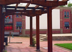 An outdoor space with wooden pergolas framing a courtyard. The ground is paved with bricks, and a mural depicting people in windows is painted on a building facade in the background. Vibrant colors on fabric decorations are visible alongside red brick walls.