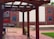 An outdoor space with wooden pergolas framing a courtyard. The ground is paved with bricks, and a mural depicting people in windows is painted on a building facade in the background. Vibrant colors on fabric decorations are visible alongside red brick walls.