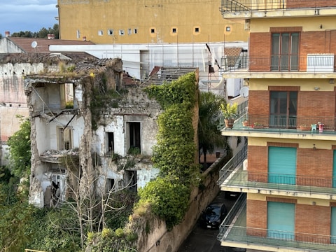 A dilapidated, overgrown building with crumbling walls stands next to a well-maintained modern apartment structure with balconies. Vegetation and vines cover parts of the ruined building, indicating neglect and nature reclaiming the space. The modern building on the right features clean lines and several balconies decorated with potted plants.