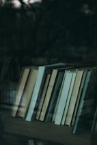 A collection of books neatly arranged on a shelf, reflected slightly in a glass surface. The background appears dark and out of focus, suggesting the image might be taken through a window or in a dimly lit room.
