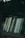 A collection of books neatly arranged on a shelf, reflected slightly in a glass surface. The background appears dark and out of focus, suggesting the image might be taken through a window or in a dimly lit room.