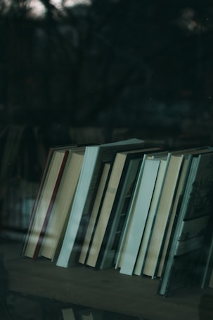A collection of books neatly arranged on a shelf, reflected slightly in a glass surface. The background appears dark and out of focus, suggesting the image might be taken through a window or in a dimly lit room.