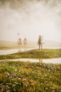 An astronaut in a spacesuit stands on a grassy field, facing away. In the distance, two figures on horseback with flags appear through the mist, creating a mysterious and surreal scene.