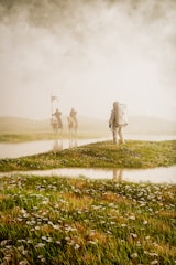 An astronaut in a spacesuit stands on a grassy field, facing away. In the distance, two figures on horseback with flags appear through the mist, creating a mysterious and surreal scene.
