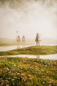An astronaut in a spacesuit stands on a grassy field, facing away. In the distance, two figures on horseback with flags appear through the mist, creating a mysterious and surreal scene.
