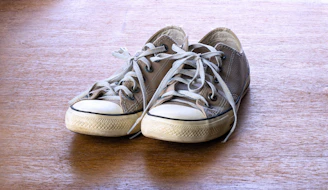 A pair of worn-out, beige canvas sneakers with white rubber soles and laces, placed side by side on a textured wooden surface.