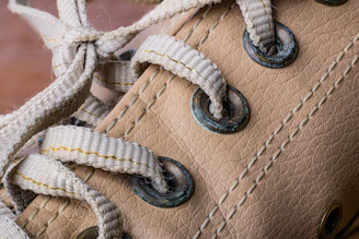 Close-up of a beige leather shoe featuring two rows of metal eyelets with white, slightly worn laces threaded through them. The eyelets show signs of rust or oxidation, adding a vintage feel.