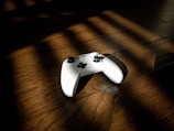 A white video game controller is resting on a dark wooden floor. Sunlight casts diagonal shadow patterns across the floor and partially on the controller, creating a play of light and shadow.