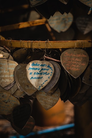 Several heart-shaped plaques with handwritten messages hang closely together. The writing is in various colors, primarily blue and red. They are suspended by a wire, creating a clustered arrangement. The lighting is dim, giving a warm, sepia-toned effect.