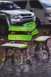 A pair of uniquely designed wooden chairs and a table, painted with vibrant green accents, are situated on a cobblestone surface. In the background, two parked vehicles, a silver SUV and a white van, are partially visible.