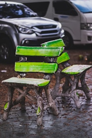 A pair of uniquely designed wooden chairs and a table, painted with vibrant green accents, are situated on a cobblestone surface. In the background, two parked vehicles, a silver SUV and a white van, are partially visible.