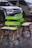 A pair of uniquely designed wooden chairs and a table, painted with vibrant green accents, are situated on a cobblestone surface. In the background, two parked vehicles, a silver SUV and a white van, are partially visible.