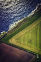 Aerial view of a coastline with clear differentiation between land and sea. The land includes geometric agricultural fields in varying shades of green and brown, with a tractor visible in one of the fields. The ocean is dark blue with waves crashing against the shore.