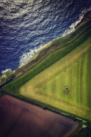 Aerial view of a coastline with clear differentiation between land and sea. The land includes geometric agricultural fields in varying shades of green and brown, with a tractor visible in one of the fields. The ocean is dark blue with waves crashing against the shore.