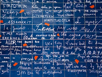 A wall covered in multiple scripts and handwritings, featuring the phrase 'I love you' written in various languages across neatly arranged blue tiles. Random red-orange shapes sporadically accent the grid-like pattern.