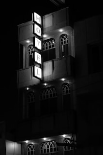 A multi-story building is highlighted by a vertical neon sign spelling 'HOTEL'. The structure features arched windows and is lit by spotlights, creating a stark contrast against the night sky.