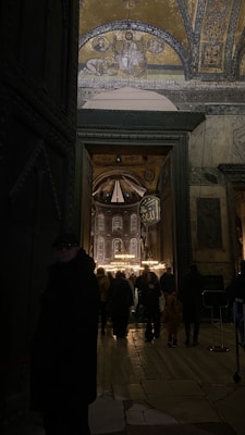 A dimly lit interior of a historic building with ornate mosaic artwork on the ceiling. A large arched doorway opens to a crowded area, where people gather under a grand chandelier. The architectural details include intricate patterns and religious symbols.