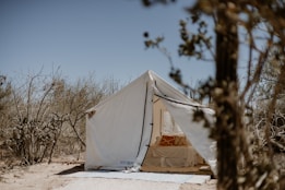 A canvas tent is set up in a dry, sparse desert environment with brush and cacti surrounding it. Inside the tent, a bed is visible with an orange and white patterned pillow. The sky above is clear and the overall setting conveys a sense of rustic outdoor living.