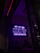 A neon sign in bright purple lettering with the text 'Biz bu işin keyif kısmındayız' is mounted on the wall. Below the sign, two people are seated together in a dimly-lit setting, suggesting a cozy atmosphere.