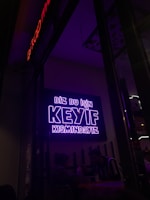 A neon sign in bright purple lettering with the text 'Biz bu işin keyif kısmındayız' is mounted on the wall. Below the sign, two people are seated together in a dimly-lit setting, suggesting a cozy atmosphere.