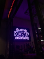 A neon sign in bright purple lettering with the text 'Biz bu işin keyif kısmındayız' is mounted on the wall. Below the sign, two people are seated together in a dimly-lit setting, suggesting a cozy atmosphere.