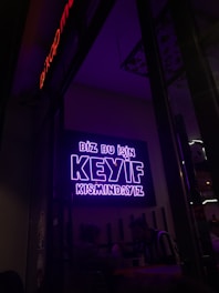 A neon sign in bright purple lettering with the text 'Biz bu işin keyif kısmındayız' is mounted on the wall. Below the sign, two people are seated together in a dimly-lit setting, suggesting a cozy atmosphere.