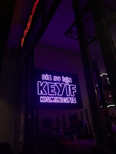 A neon sign in bright purple lettering with the text 'Biz bu işin keyif kısmındayız' is mounted on the wall. Below the sign, two people are seated together in a dimly-lit setting, suggesting a cozy atmosphere.