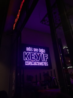A neon sign in bright purple lettering with the text 'Biz bu işin keyif kısmındayız' is mounted on the wall. Below the sign, two people are seated together in a dimly-lit setting, suggesting a cozy atmosphere.