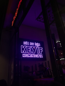 A neon sign in bright purple lettering with the text 'Biz bu işin keyif kısmındayız' is mounted on the wall. Below the sign, two people are seated together in a dimly-lit setting, suggesting a cozy atmosphere.