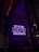 A neon sign in bright purple lettering with the text 'Biz bu işin keyif kısmındayız' is mounted on the wall. Below the sign, two people are seated together in a dimly-lit setting, suggesting a cozy atmosphere.