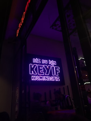 A neon sign in bright purple lettering with the text 'Biz bu işin keyif kısmındayız' is mounted on the wall. Below the sign, two people are seated together in a dimly-lit setting, suggesting a cozy atmosphere.