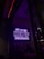 A neon sign in bright purple lettering with the text 'Biz bu işin keyif kısmındayız' is mounted on the wall. Below the sign, two people are seated together in a dimly-lit setting, suggesting a cozy atmosphere.